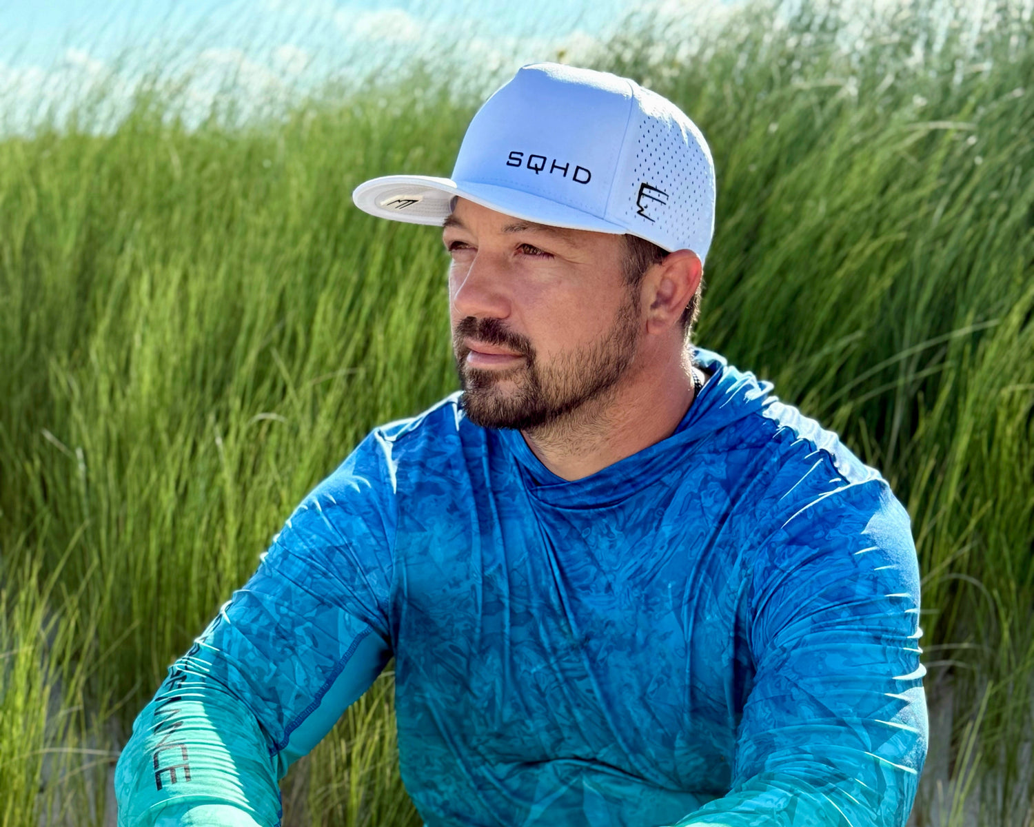 Man wearing a blue long-sleeve shirt and cap sitting on sand with grass in the background